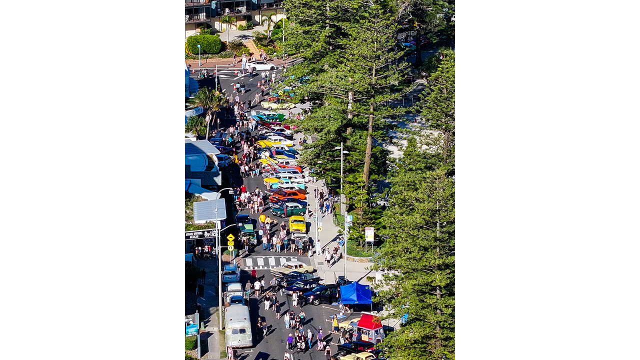 An aerial view of the street filled with people and classic and custom cars on display, with beautiful green trees lining the street on the right.