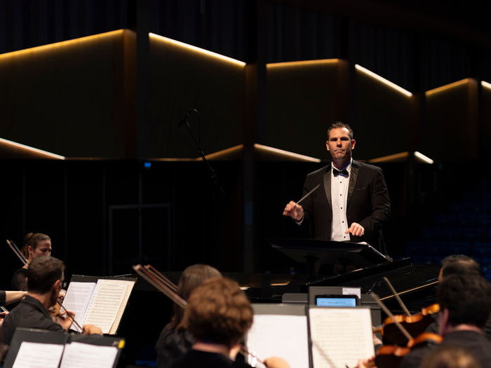 A conductor stands on a podium, illuminated by stage lights, energetically leading an orchestra with a baton, surrounded by musicians playing various instruments.