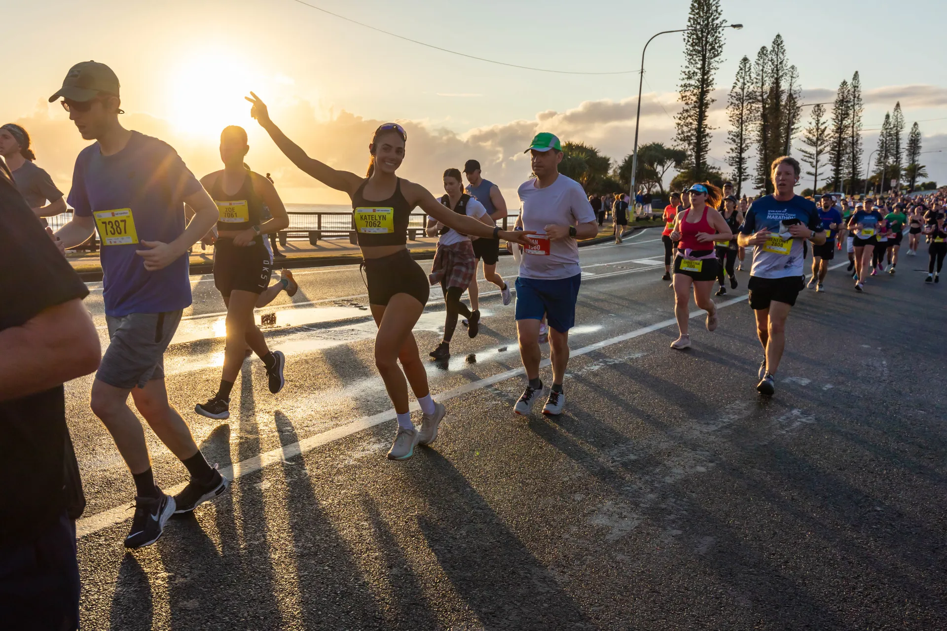 An early morning photo featuring 8 runners in the main area and hundreds of others out of focus behind them. One female runner has her arms stretched out, acknowledging the photographer. The rising sun appears in the background adding a golden glow to the photo.