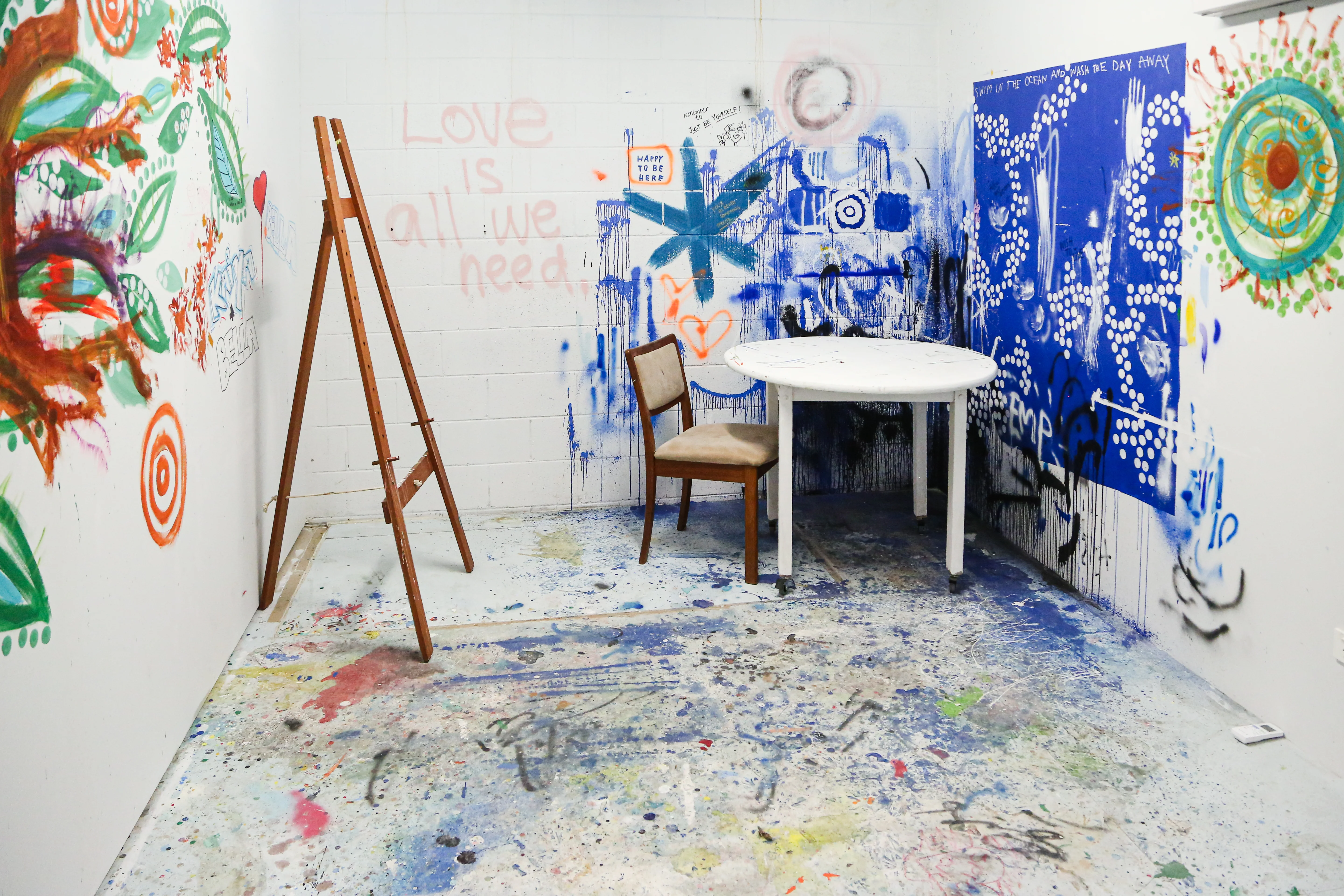 Artist space with paint on the floor, an easel, table and chair, and artworks painted directly onto the white walls