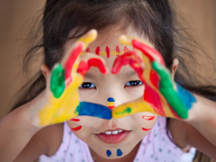 Young girl with colourful paint on her hands making a shape of a heart with her hands.