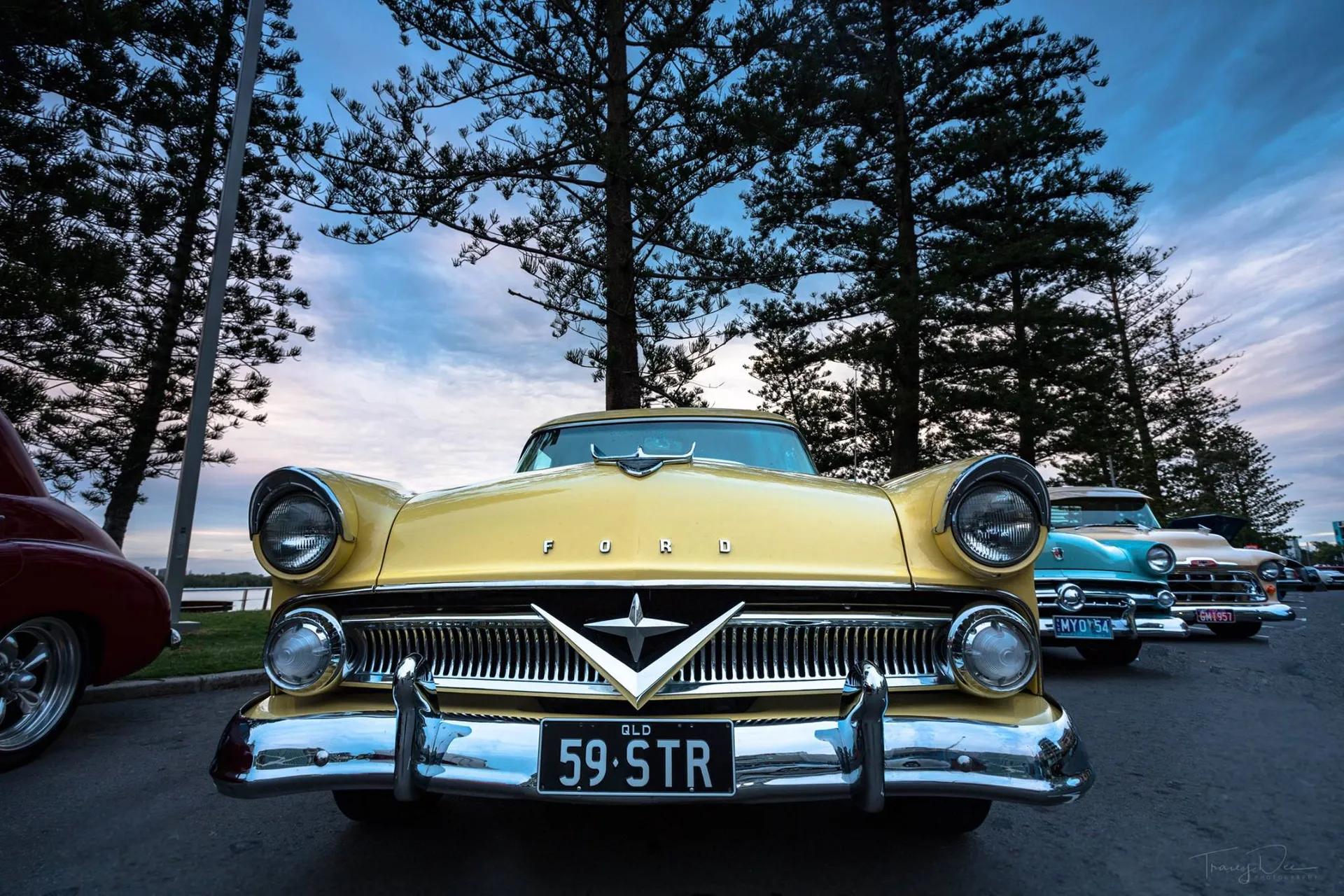 The front of a 1959 Ford car - yellow in colour. Trees in the background and similar cars to the right.