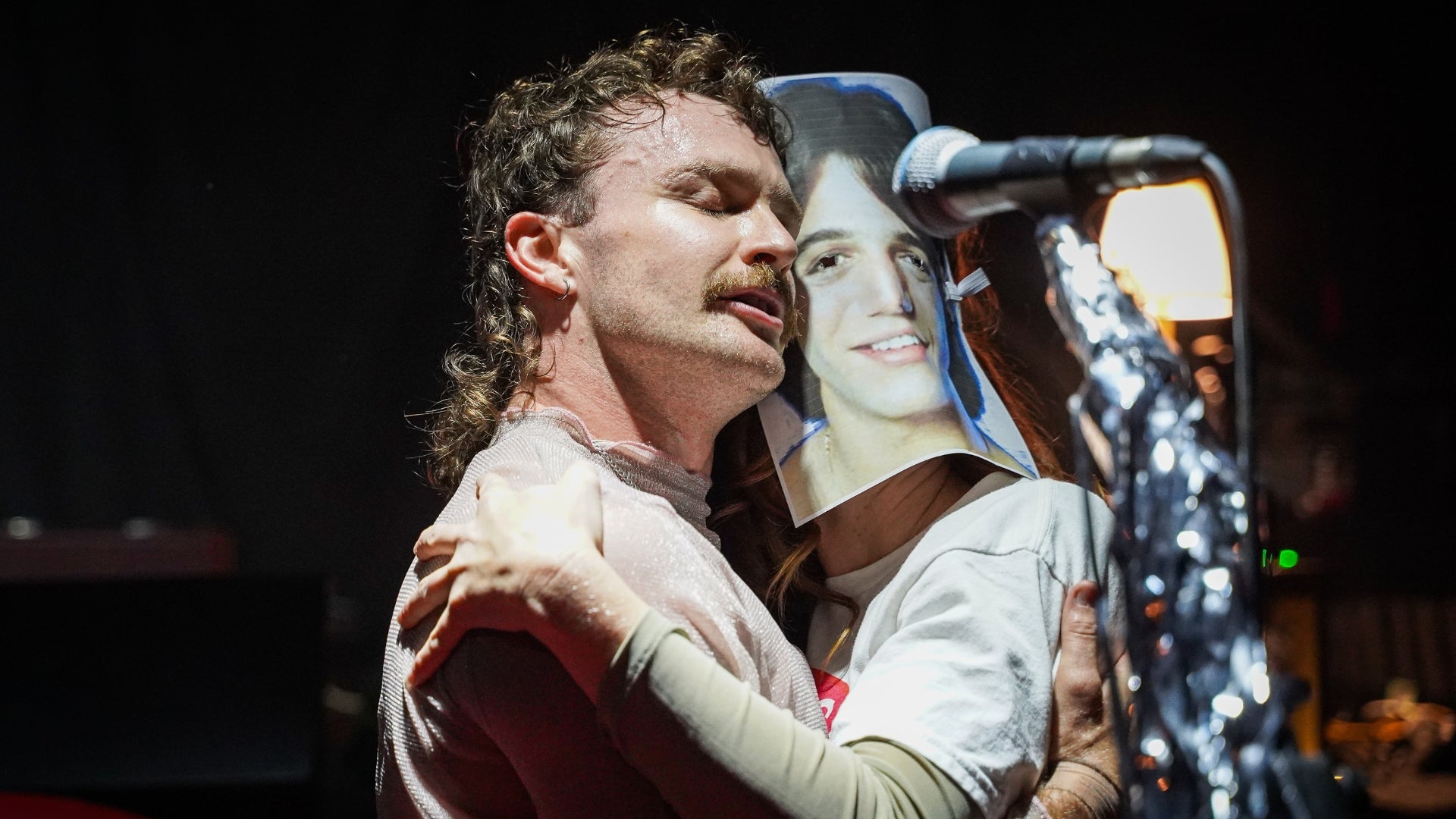 Two people standing in front of a microphone, hugging. One person has a moustache, a mullet hairstyle and their eyes are closed. The other is wearing a mask, featuring the face of Tony Danza.