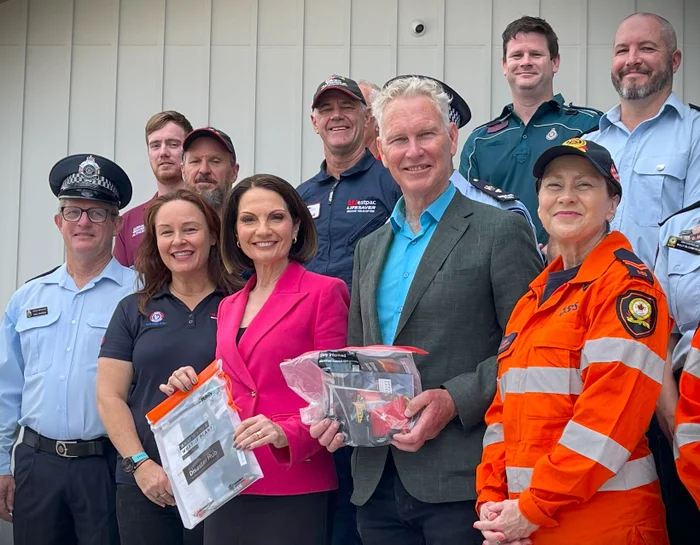Sunshine Coast Mayor Natoli and Noosa Mayor Wilkie hold emergency packs and look at the camera. They are surrounded by Emergency Service Personnel in uniform.