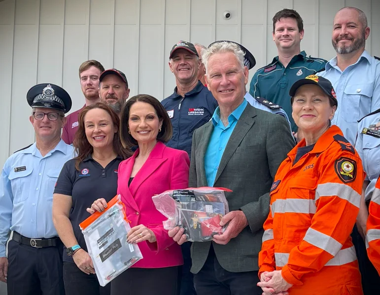 Sunshine Coast Mayor Natoli and Noosa Mayor Wilkie hold emergency packs and look at the camera. They are surrounded by Emergency Service Personnel in uniform.