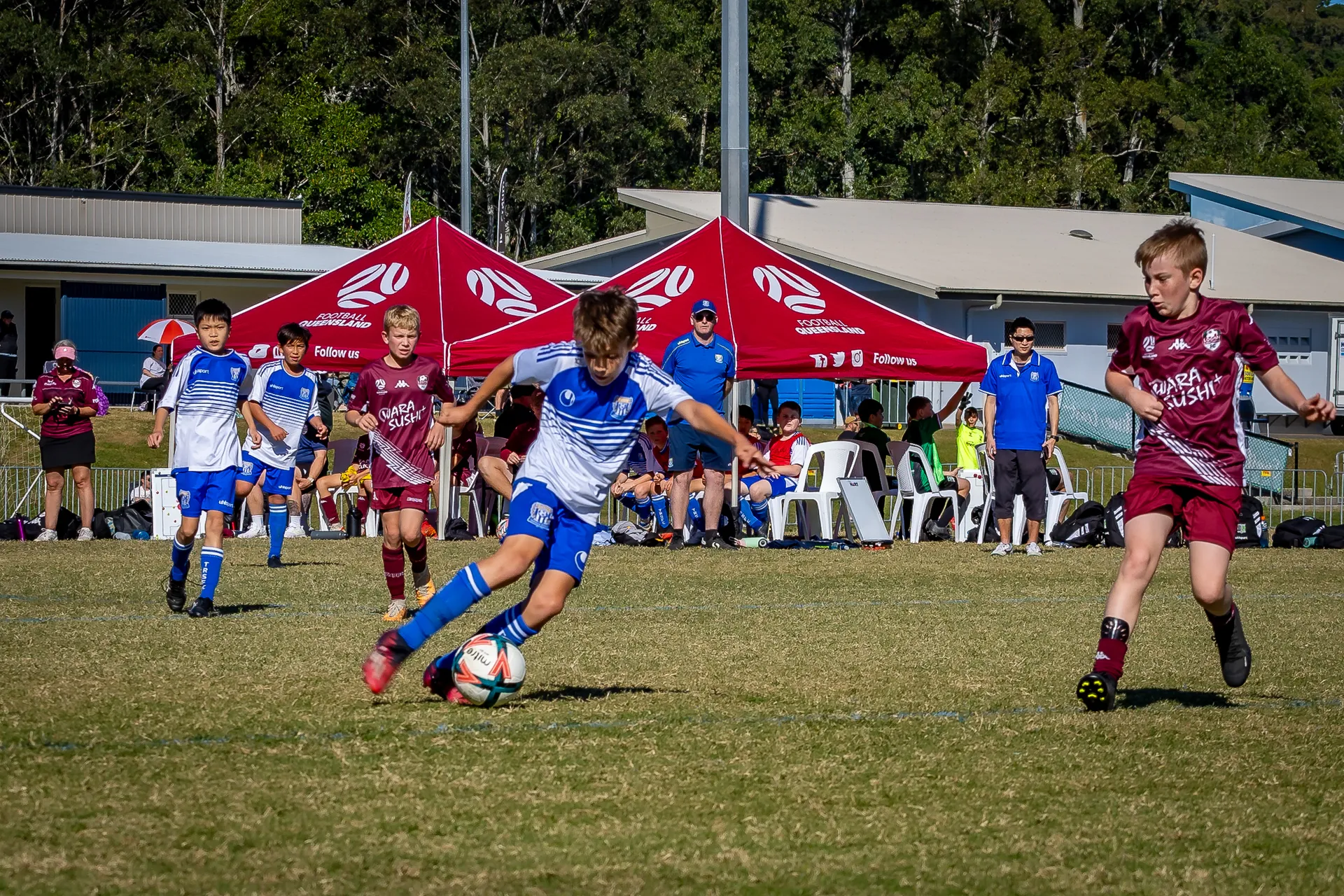 Junior football players - (centre) blue and white jersey player about to kick the ball and on the left a maroon jersey player is running towards the ball - other players behind them, as well as tents and chairs in the background.