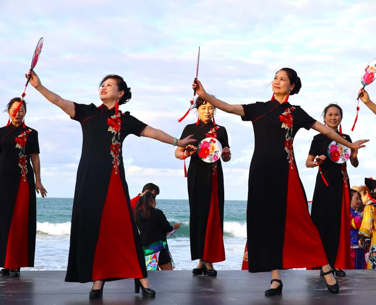 Women in traditional Chinese dresses and holding fans, stand posing on a stage with the ocean in the background.