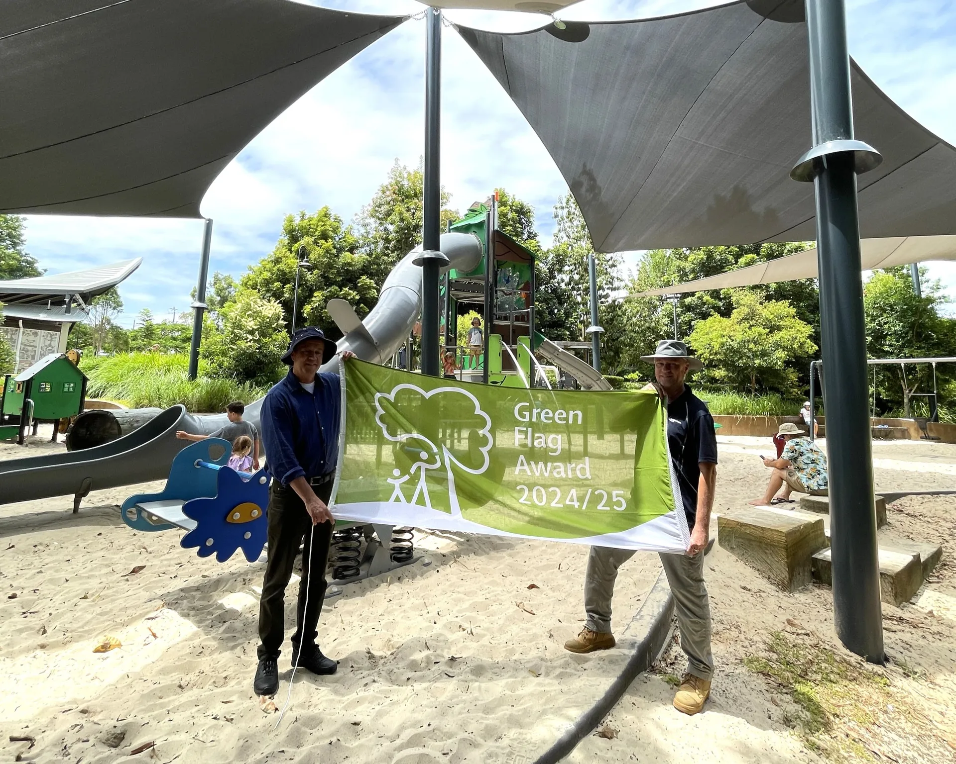 At Buderim Village Park Green. Two people standing on a sand filled playground with a slide in the background. Holding up the Green Flag Award 2024/25 Flag.