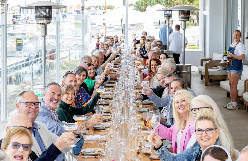 A long table setting with guests holding up glasses to cheers to the camera.