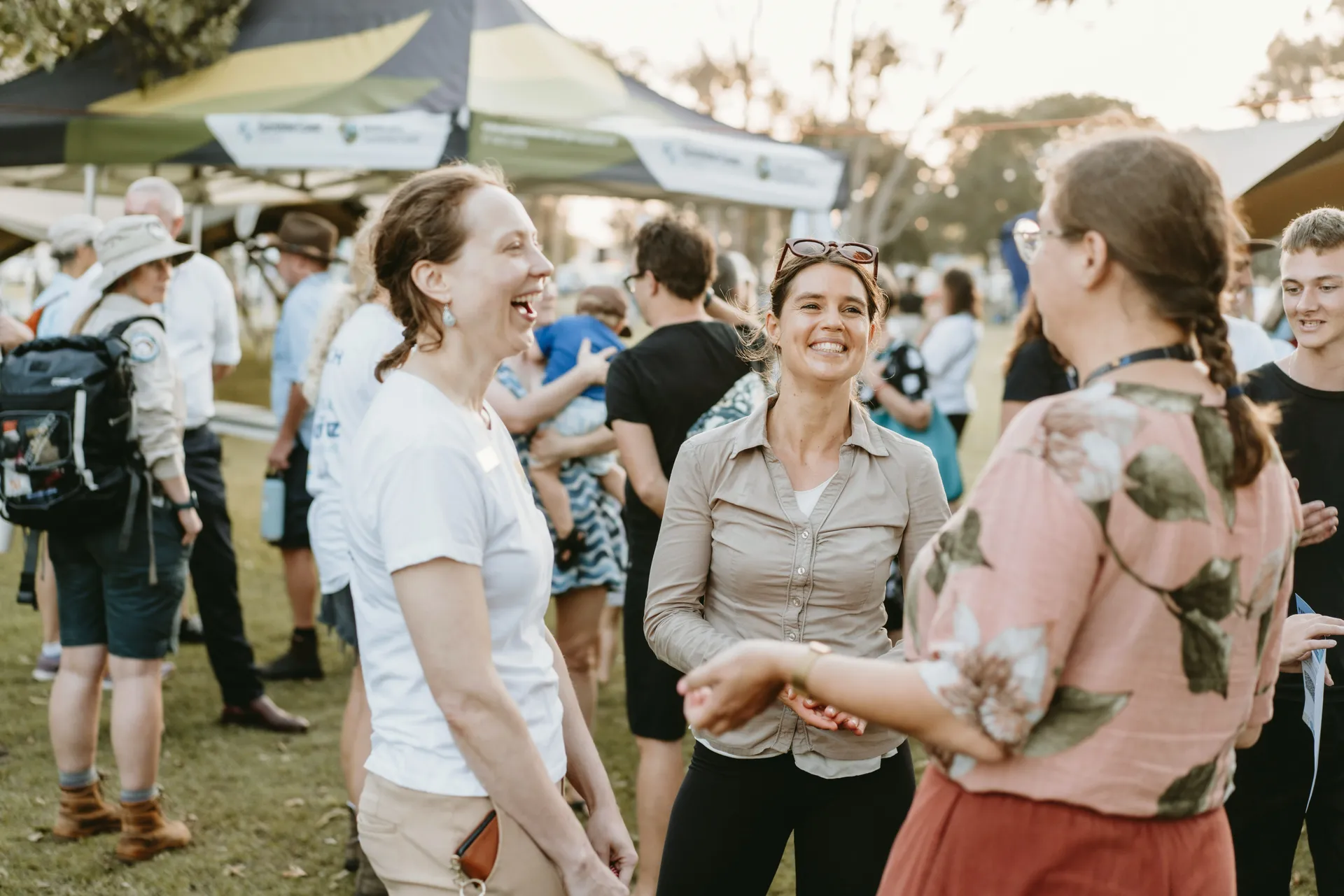 "A group of people laughing and chatting outdoors at a community event, enjoying the sunshine and each other's company."