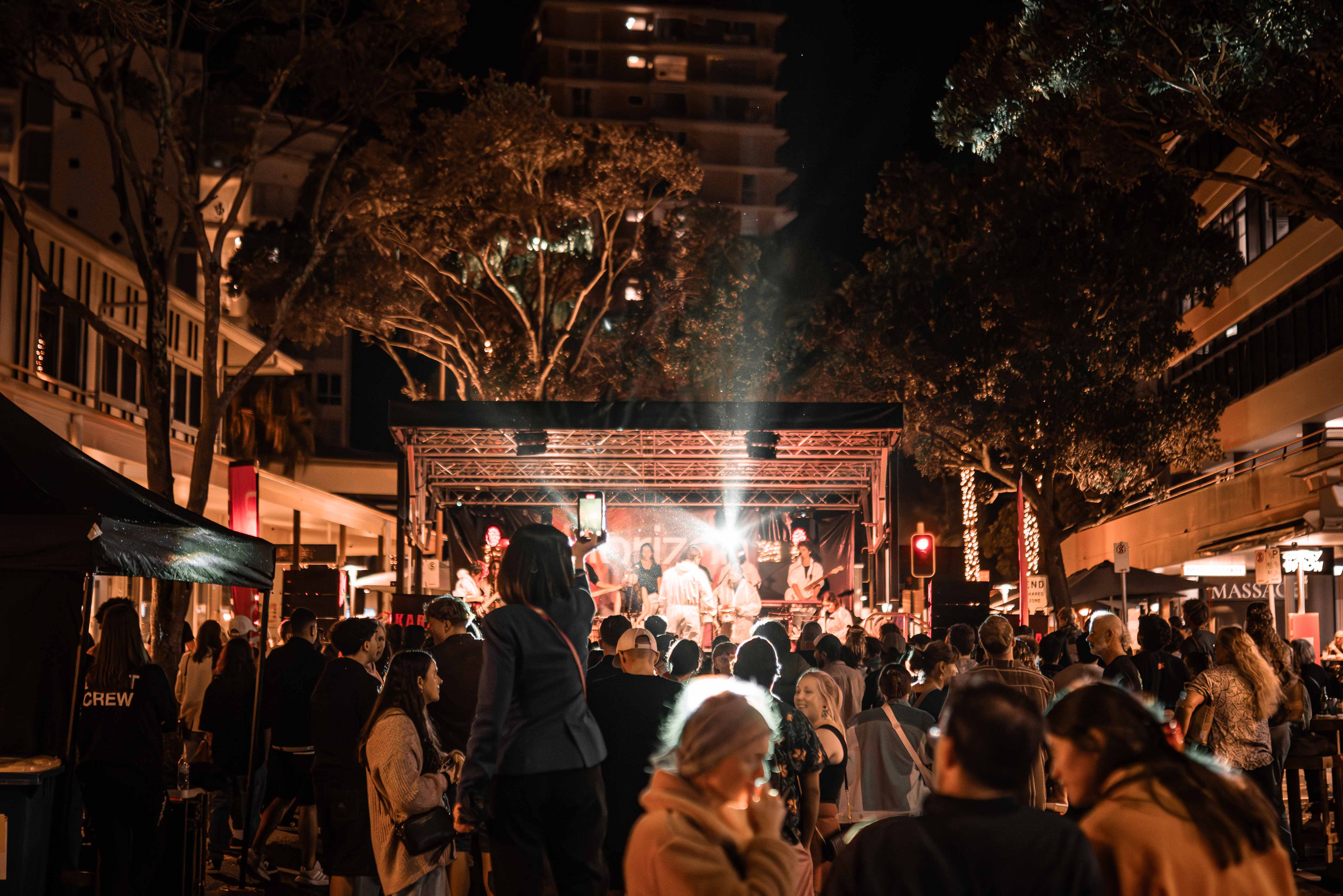 Festivel visitors in front of a stage, with trees and building above.
