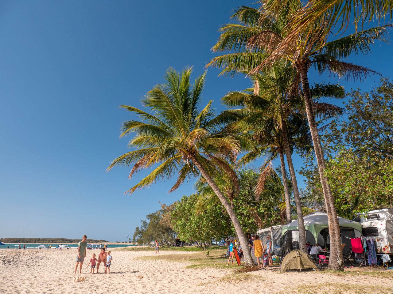 Blue sky and palm trees with tents set up underneath, a family of four on the beach playing a game.