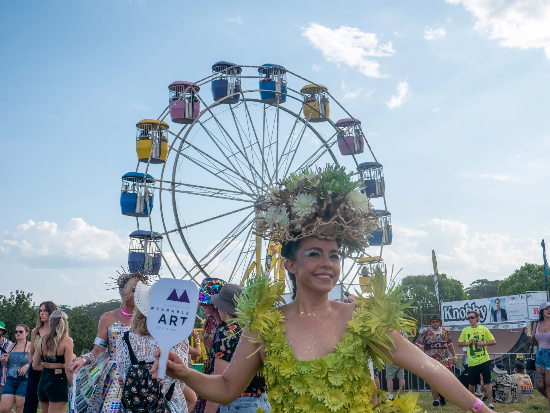 A model from the Australian Wearable Art Festival wearing a green coloured flower outfit and holding a small sign to promote the AWAF and she is standing in front of the ferris wheel at the 2024 festival.