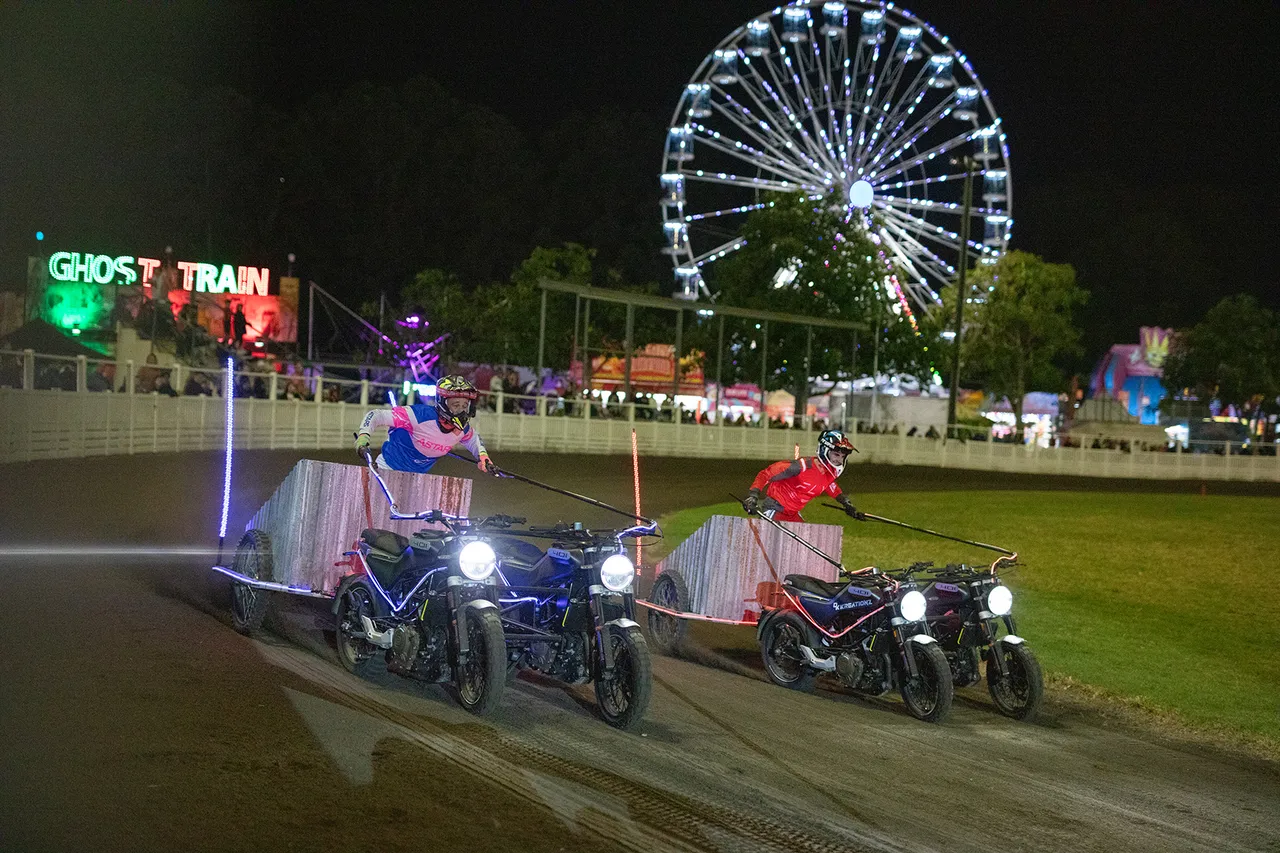 The ferris wheel lights in the background, and two people in 'chariots' with two motorcycles each pulling them around the show ring.
