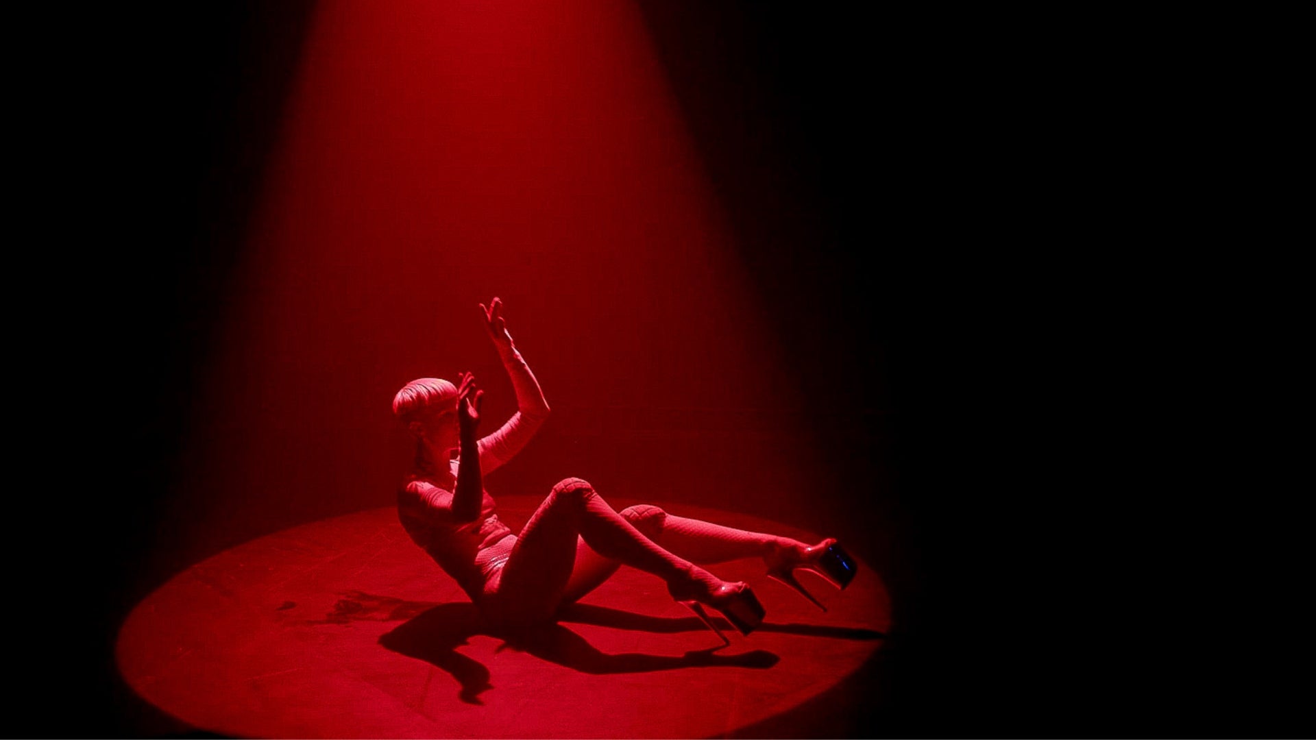 Person on small round stage, seated on backside with legs and arms positioned in air. The person is wearing high heels and a leotard. The stage is bathed in red light.