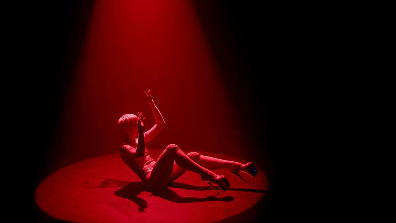 Person on small round stage, seated on backside with legs and arms positioned in air. The person is wearing high heels and a leotard. The stage is bathed in red light.