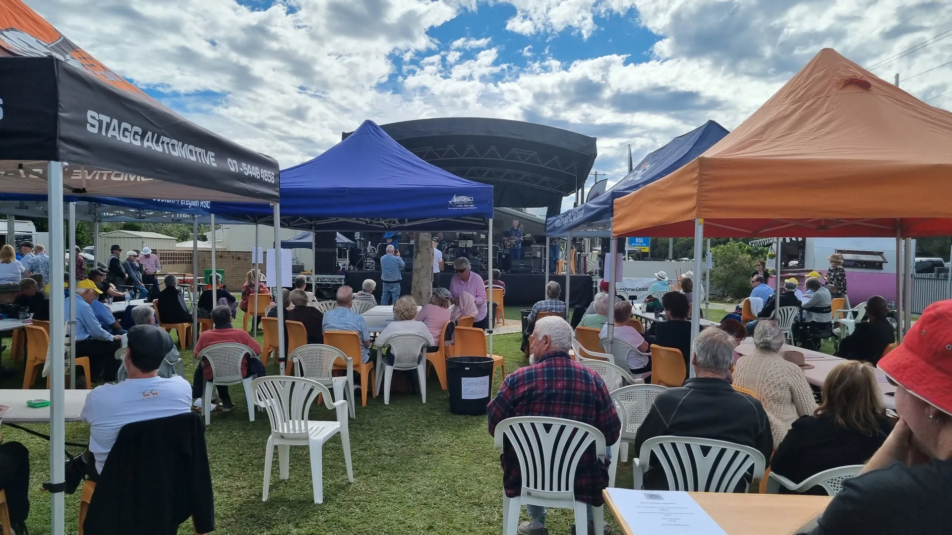 A large group of older people are seated on plastic chairs in clusters under marquees in front of a stage.