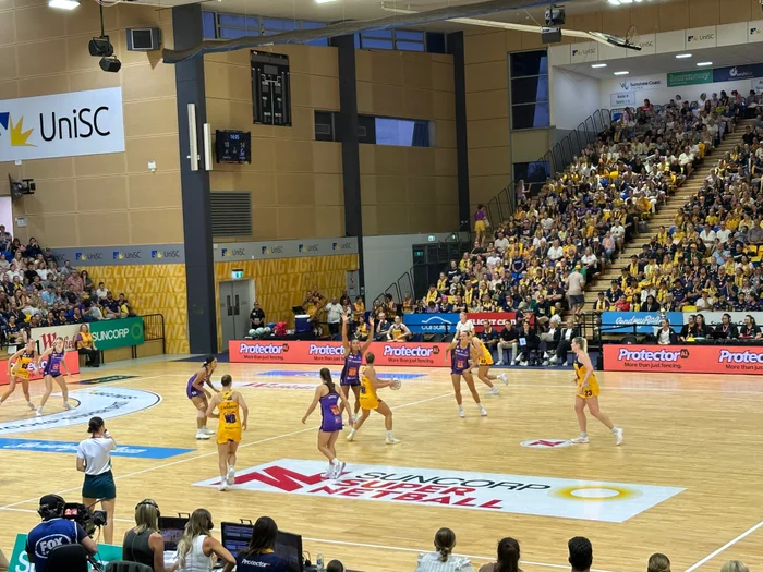 Sunshine Coast Lightning in action playing competative netball game on a netball court. Yellow and purple uniforms. 