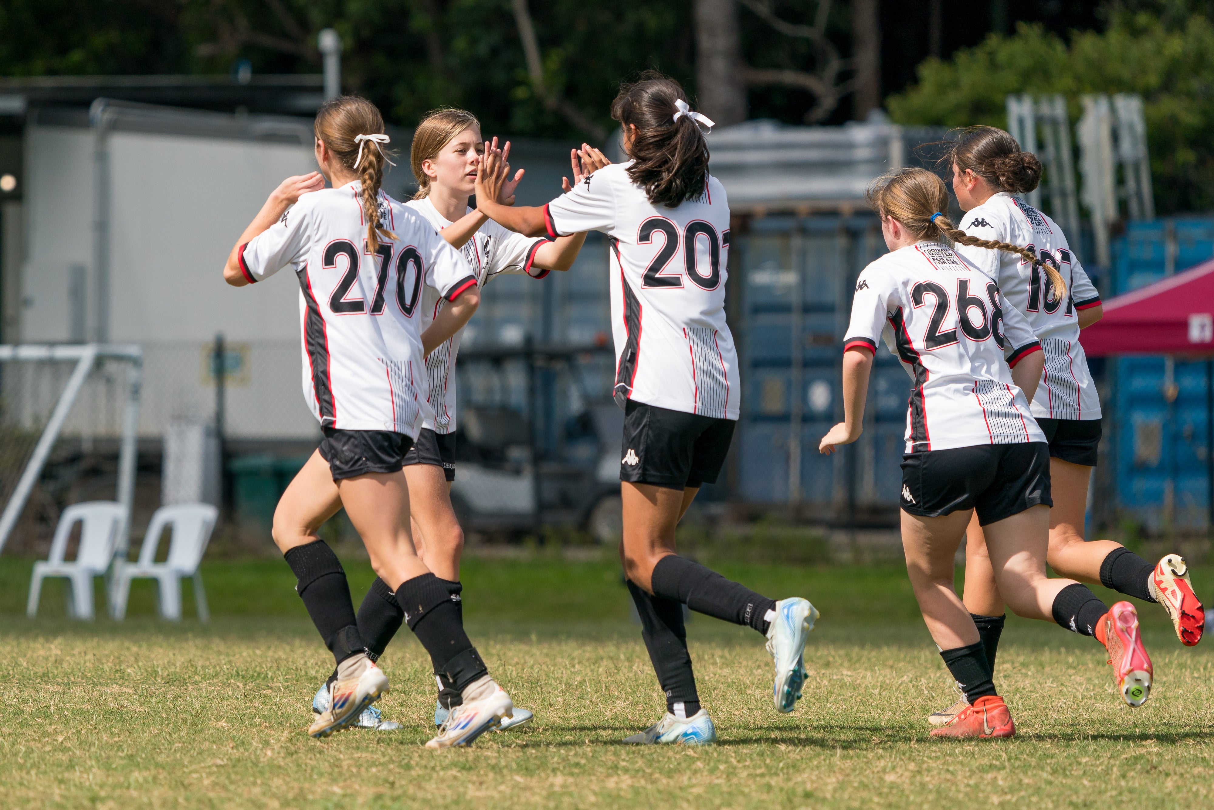 Five female players from the same team in white jerseys, black shorts, black socks to their knees, two girls are congratulating each other with both hands up for a two-handed high five.