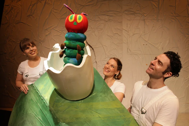 'The Very Hungry Caterpillar Show' image of a large puppet bright green with a red head and cute green and yellow eyes, popping out of a white egg sitting on a large green leaf. Theatre cast smiling up in white t-shirts at the puppet. Cast members Tina Jackson, Eleanor Stankiewicz and Christopher Vernon in The Very Hungry Caterpillar Show