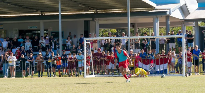 A young football (soccer) player (dressed in blue shirt and red shorts) is running towards centre field after kicking a goal, with the goalie (in yellow) lying on the ground in front of the goal netting. Lots of spectators along the fenceline in the background behind.