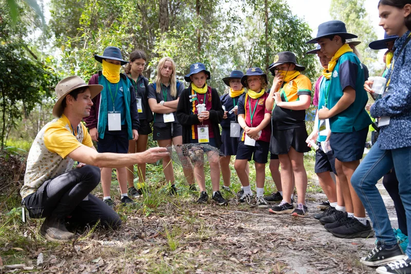 A man teaches a group of school kids about environment processes in a forest setting. 