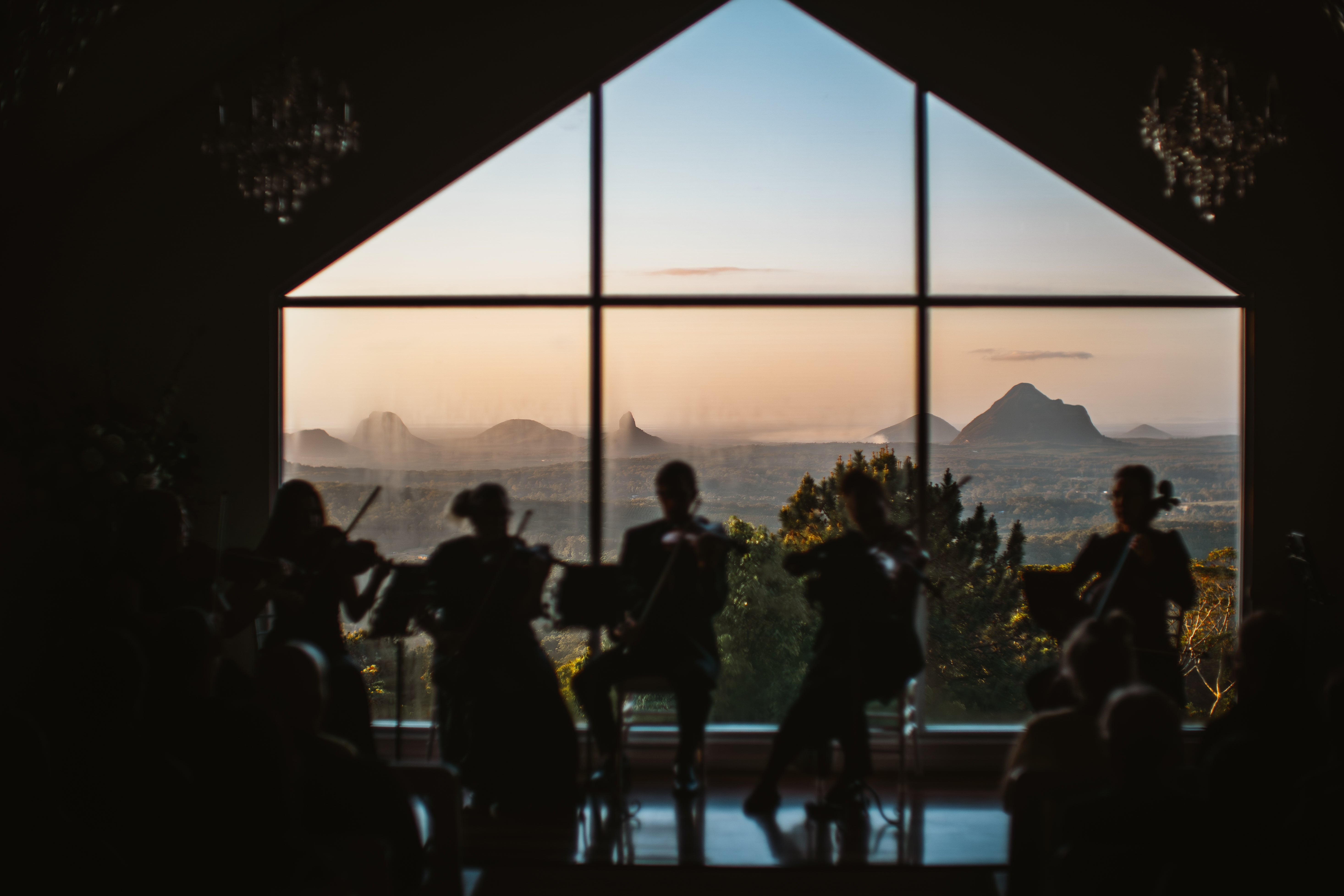A silhouette of 5 musicians sitting in front of a window, with the sky and Glasshouse Mountains in the background.