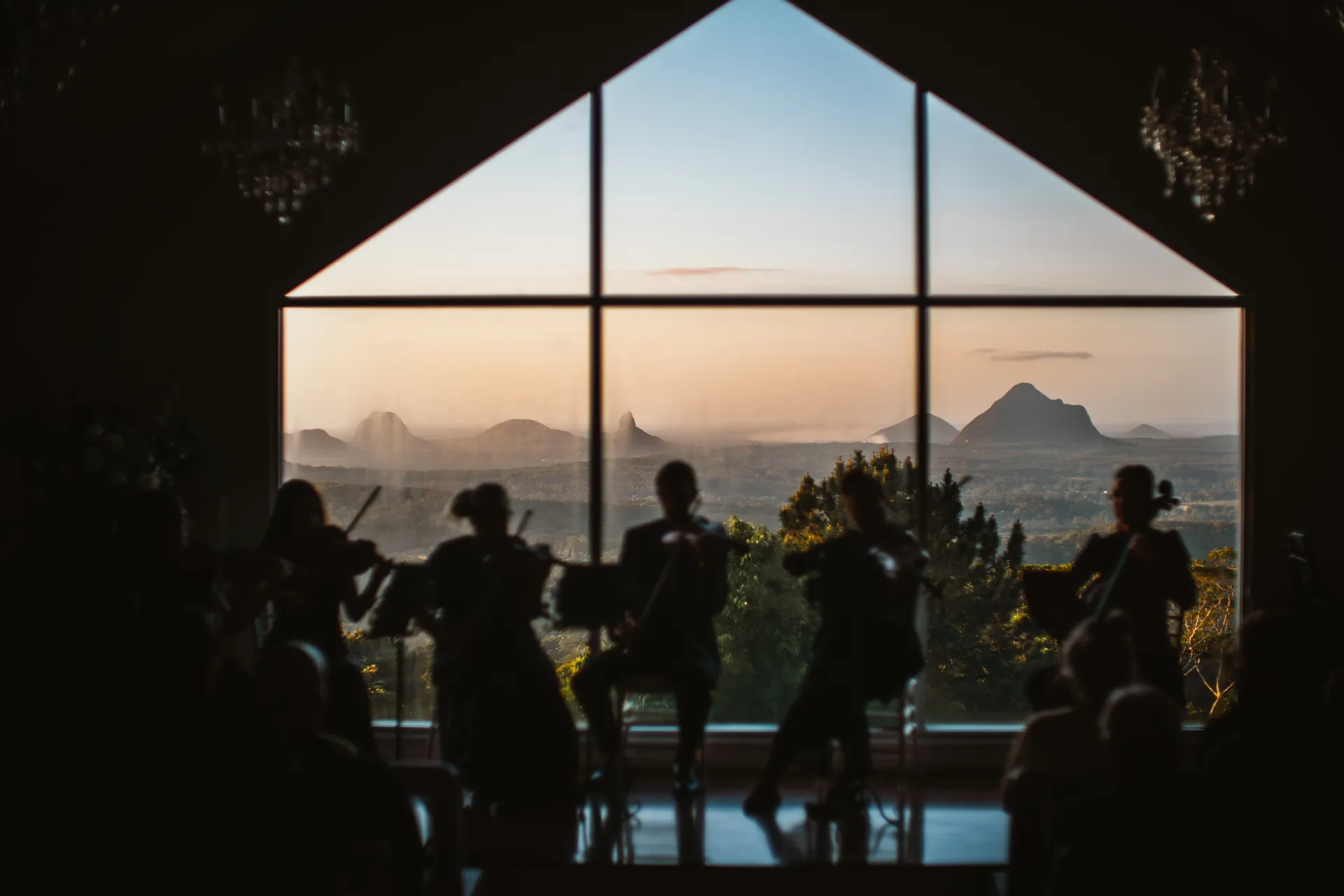 A silhouette of 5 musicians sitting in front of a window, with the sky and Glasshouse Mountains in the background.