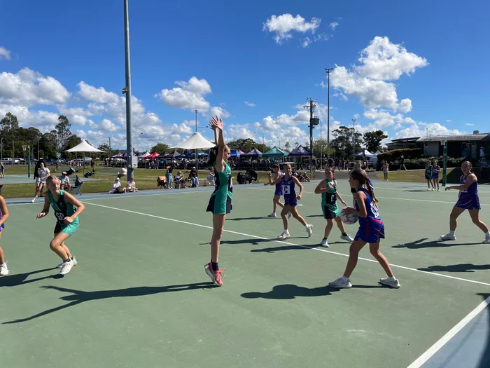 School aged netballers on the court - with one player on tippy toes trying to block a pass.