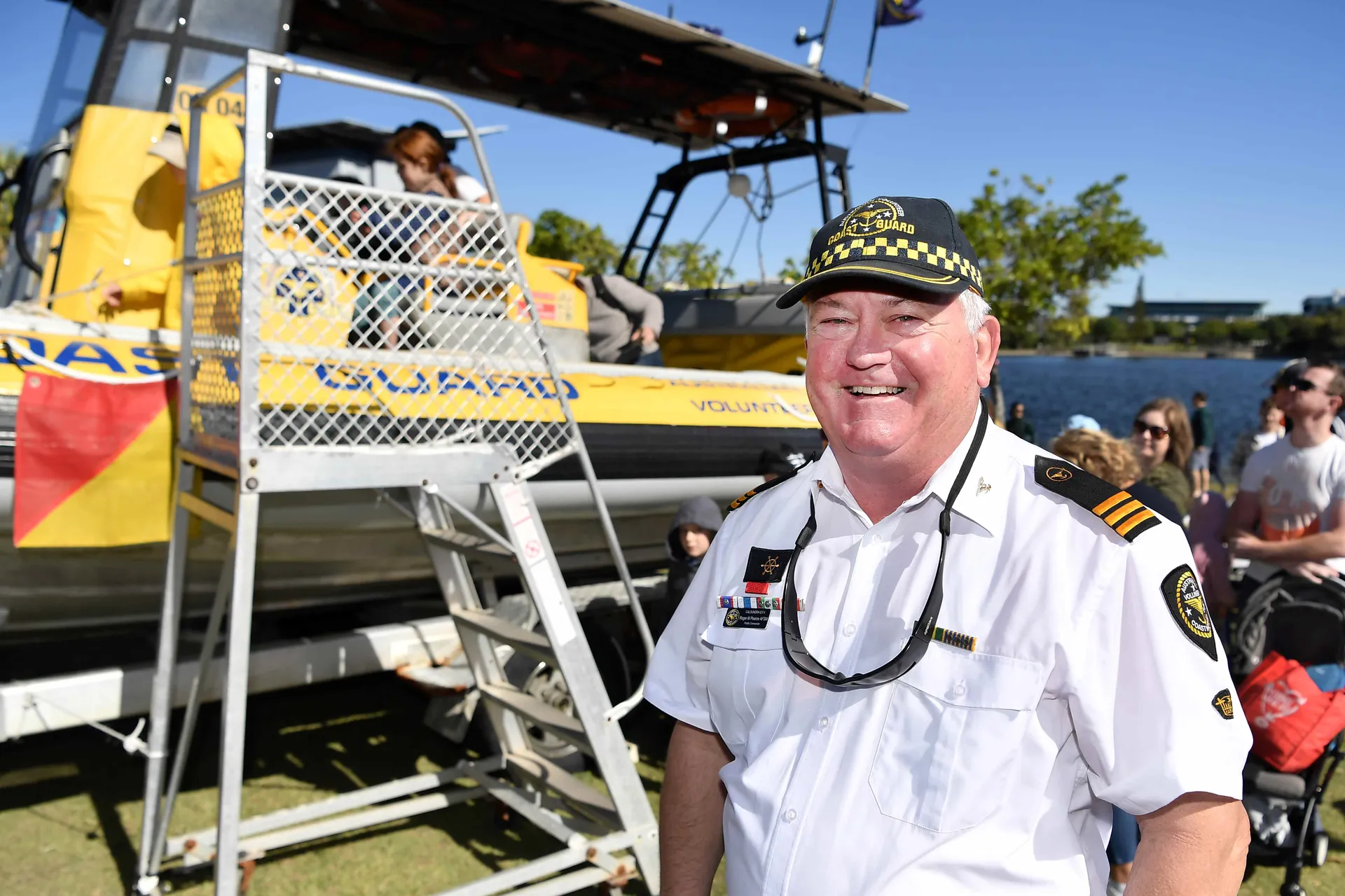 An older man in uniform stands in front of a Coast Guard boat with children and their parents in the background.