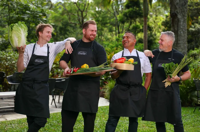 Four chefs wearing black Curated Plate Aprons standing shoulder to shoulder holding fresh local produce