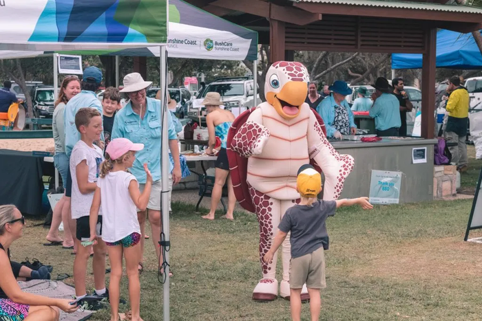 A person dressed in a turtle suit hugs a young child. In the background, a busy festival scene unfolds with people cooking on a barbecue and stall holders engaging with attendees.