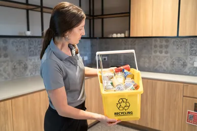 Woman in a grey shirt and black pants holds a small yellow recycle bin, full of rubbish in a kitchen.