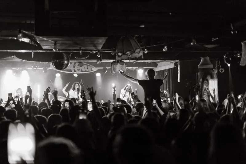 Taken from the back of the venue, this is a black and white picture of performers on stage at the Solbar and the crowd with their hands up, one person is up on someone's shoulders - higher than the rest of the crowd.