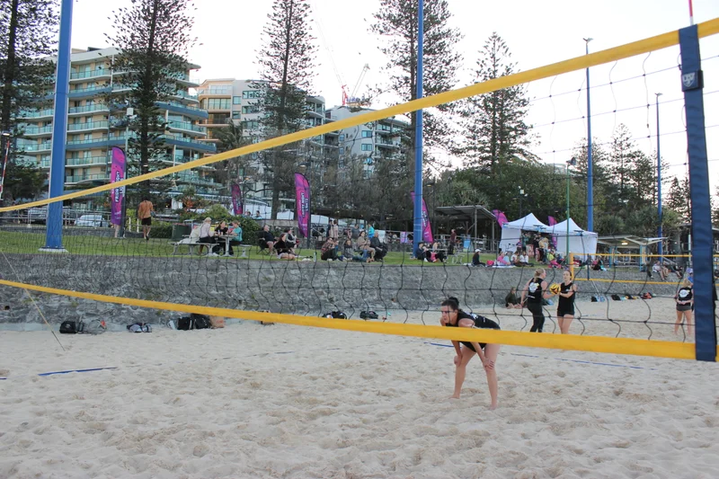 Volleyball net setup during game at Mooloolaba Beach. Woman focusing on game with crowd behind her on grassy area. 