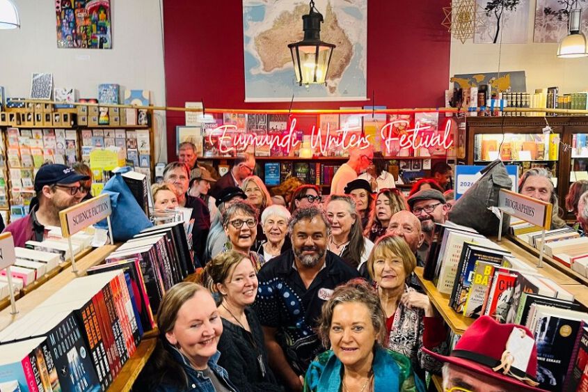 A number of people (writers) gathered in amongst bookstore shelving