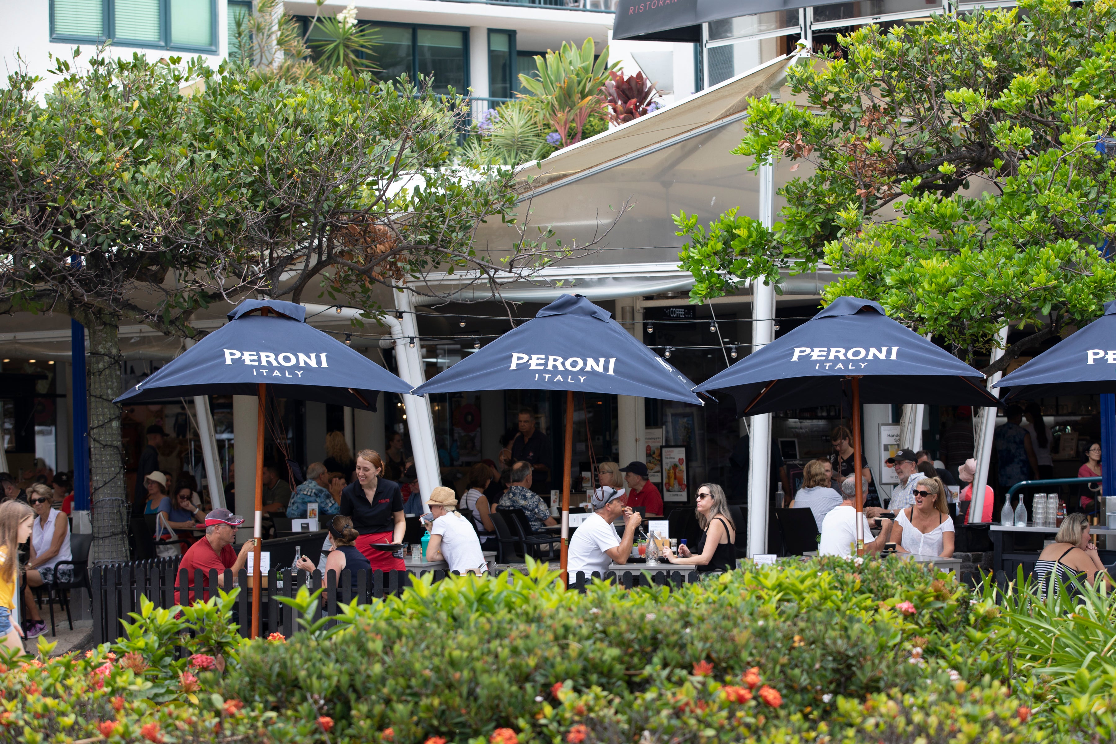 Groups of people enjoying food at a cafe under blue umberellas amongst green trees and garden bushes.