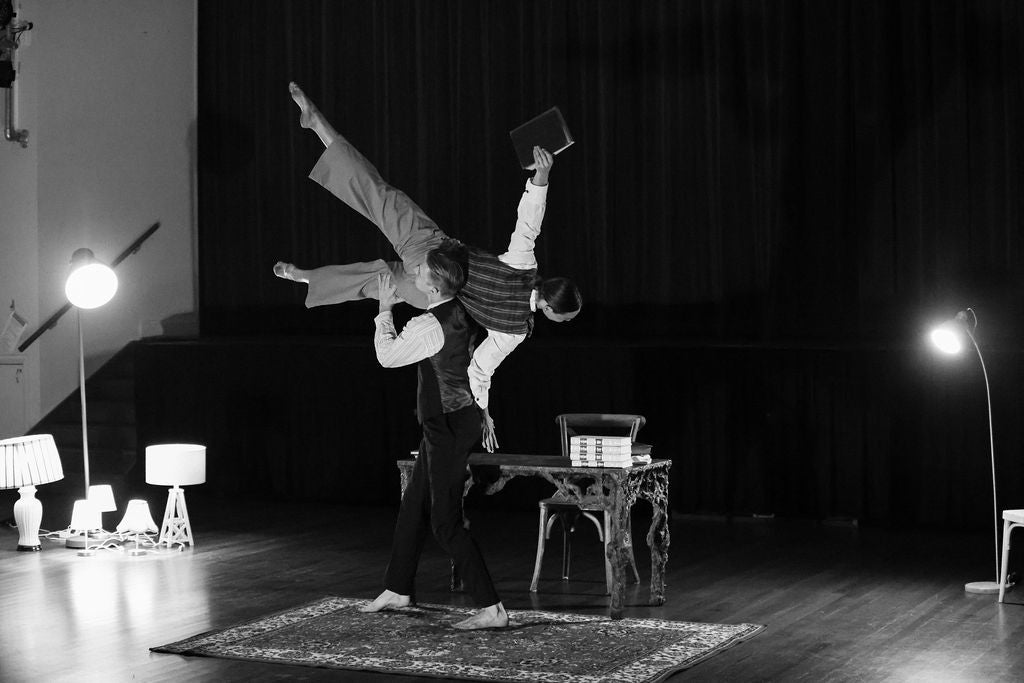 Two dance artists performing. One is holding the other over their head in a room with a table, lamps and a rug on the wooden floor