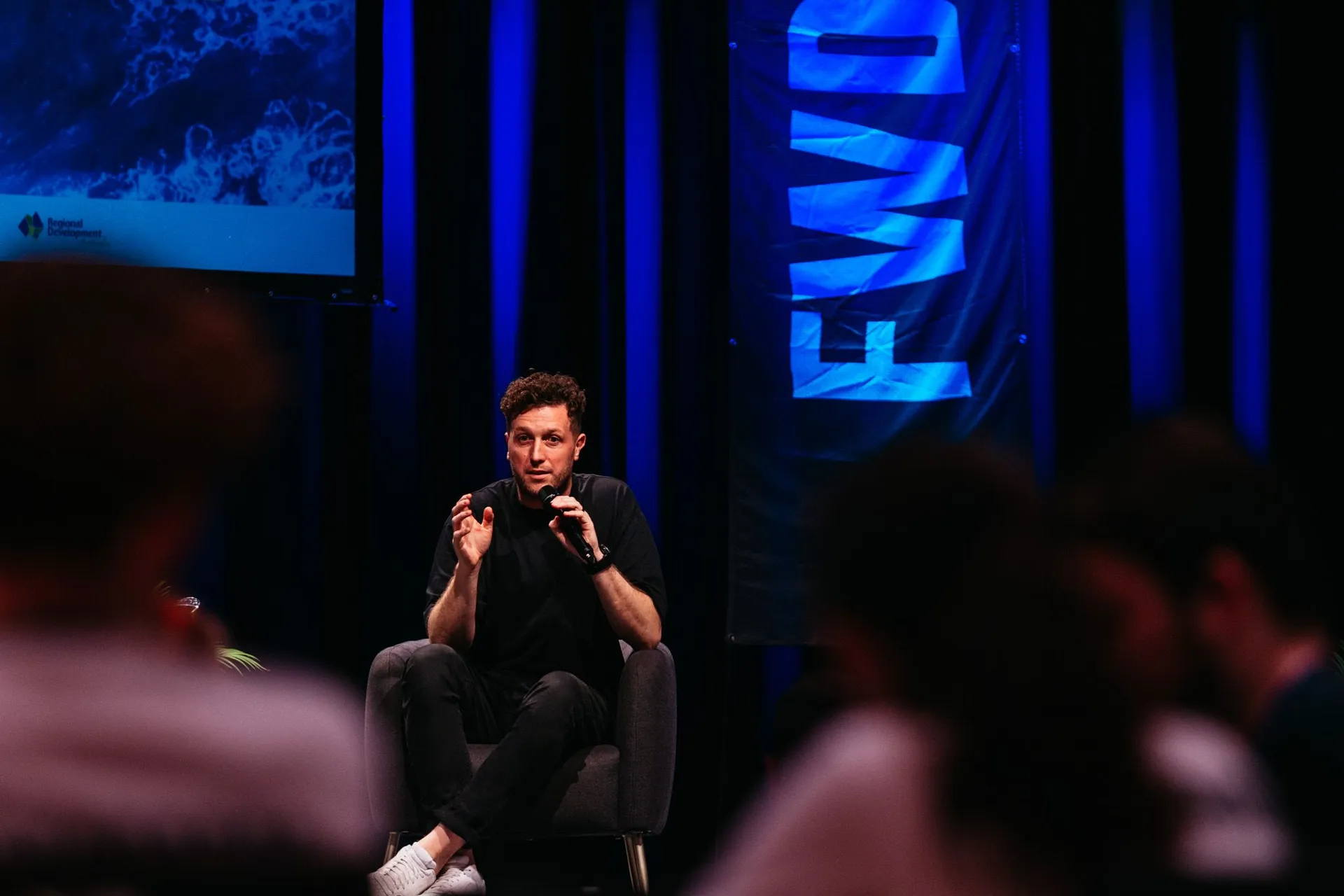 A man sits on a stage speaking intently into a microphone to an audience.