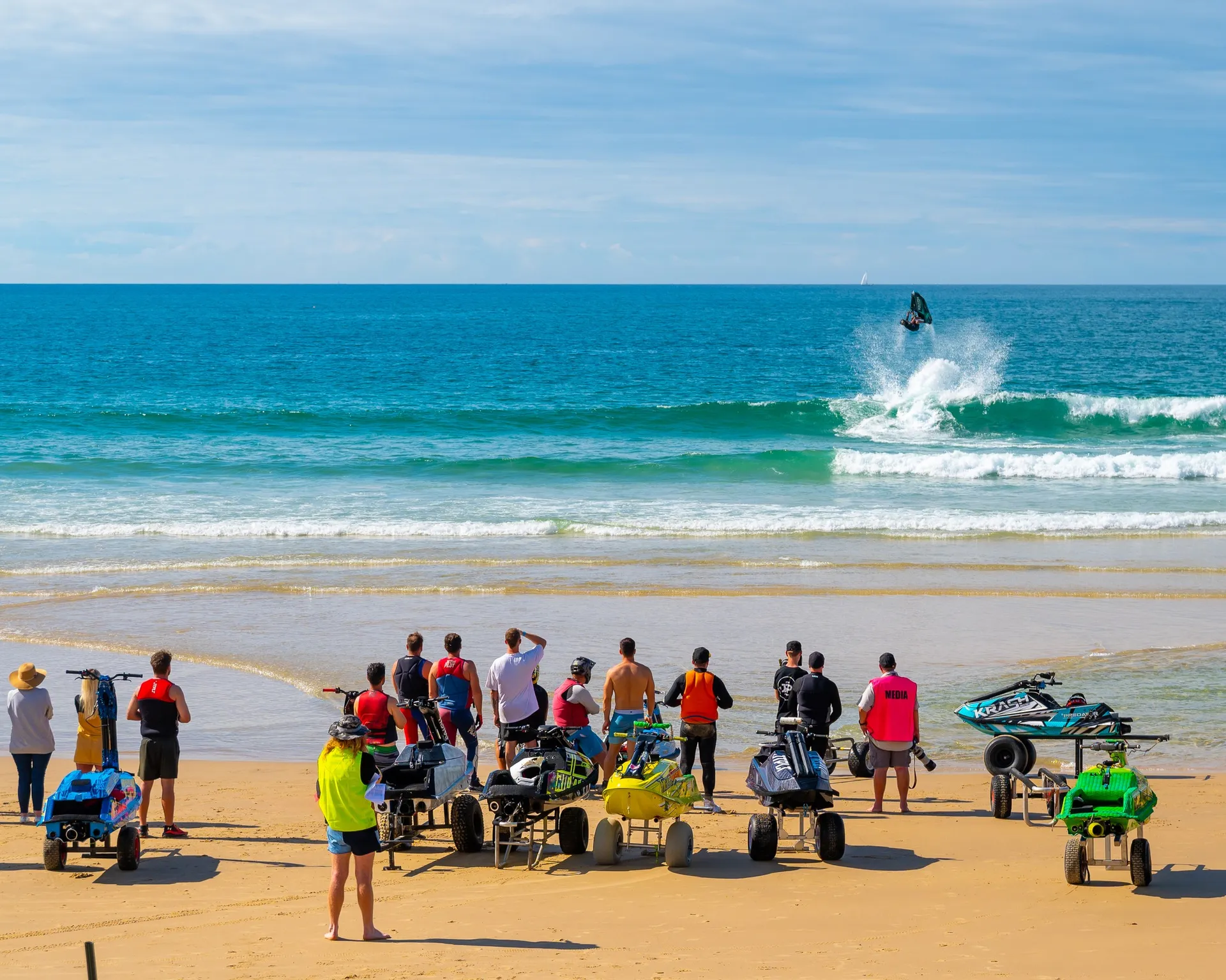 A group of people watching a jet ski rider flipping up off a wave. People are standing amongst some jetski's on trailers on the beach.