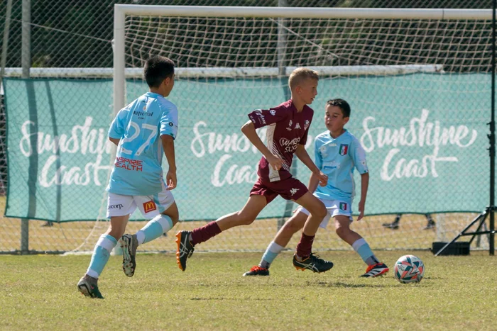 Three players on the field, two boys in blue jerseys, one boy in maroon who is racing to kick the ball, while the two in blue are trying to defend. 'Sunshine Coast' is written on signage on fencing behind them.