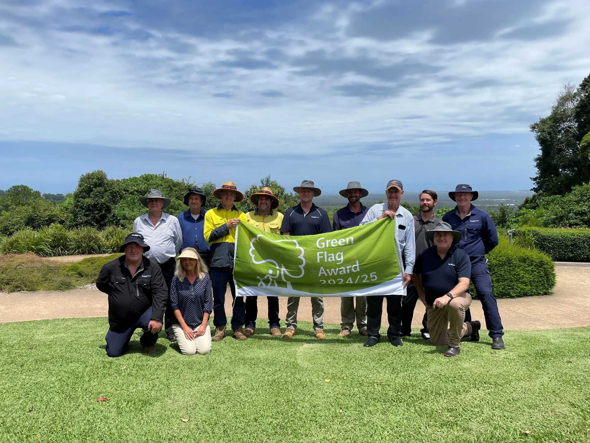 Group of twelve people all with sun protection hats. Holding the Green Flag Award flag for 2024  2025. Standing on lush green manicured land and blue skies with subtle spread of white clouds. Green bushland behind the people.