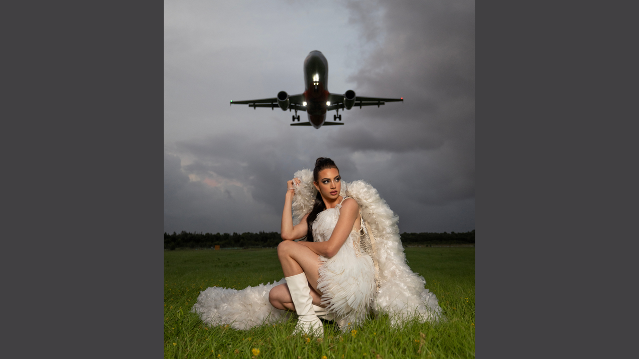 Model Jodi Urli wears Plastic Rain by Natalie Hamblin (2024 Runner-up Trashion). Jodi is poised on a grass field with an plane about to fly overhead.