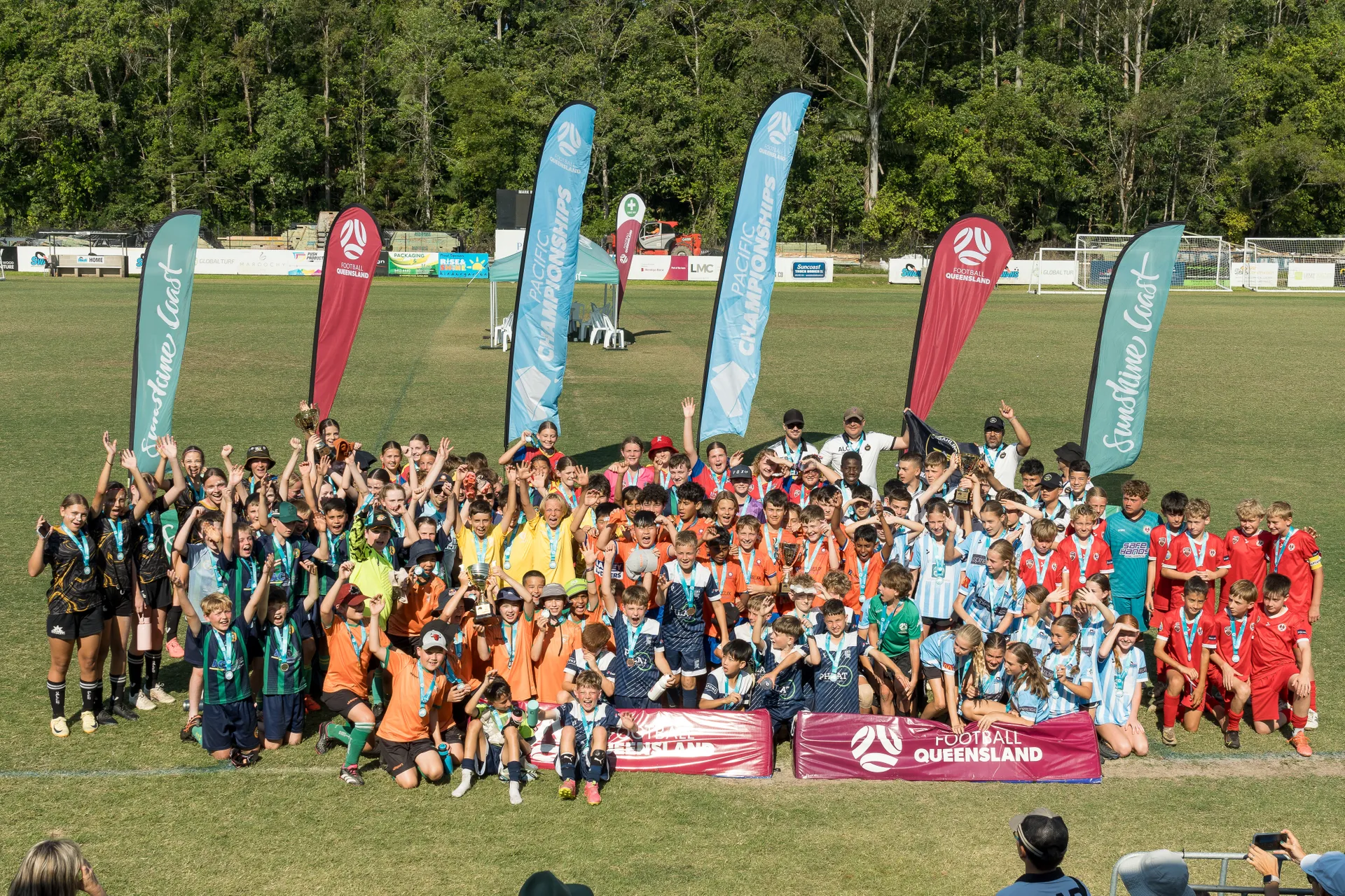 Set on a grassy green sportsground, a group picture with all players in multiple teams / colour uniforms, with a number of vertical flags in the background - Football Queensland, Pacific Championships, Sunshine Coast