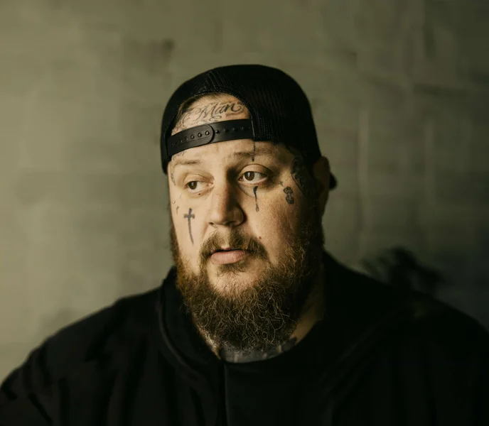 A portrait style image of performer - Jelly Roll, with a black hat (backwards) and black t-shirt