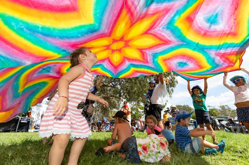 Children playing on soft green lush grass, some sitting and standing under a rainbow parachute. The fabric filled with a colourful pattern of an yellow and bright orange flower in the centre, and surrounded with bright pink, blue, green, yellow and orange pattern. 