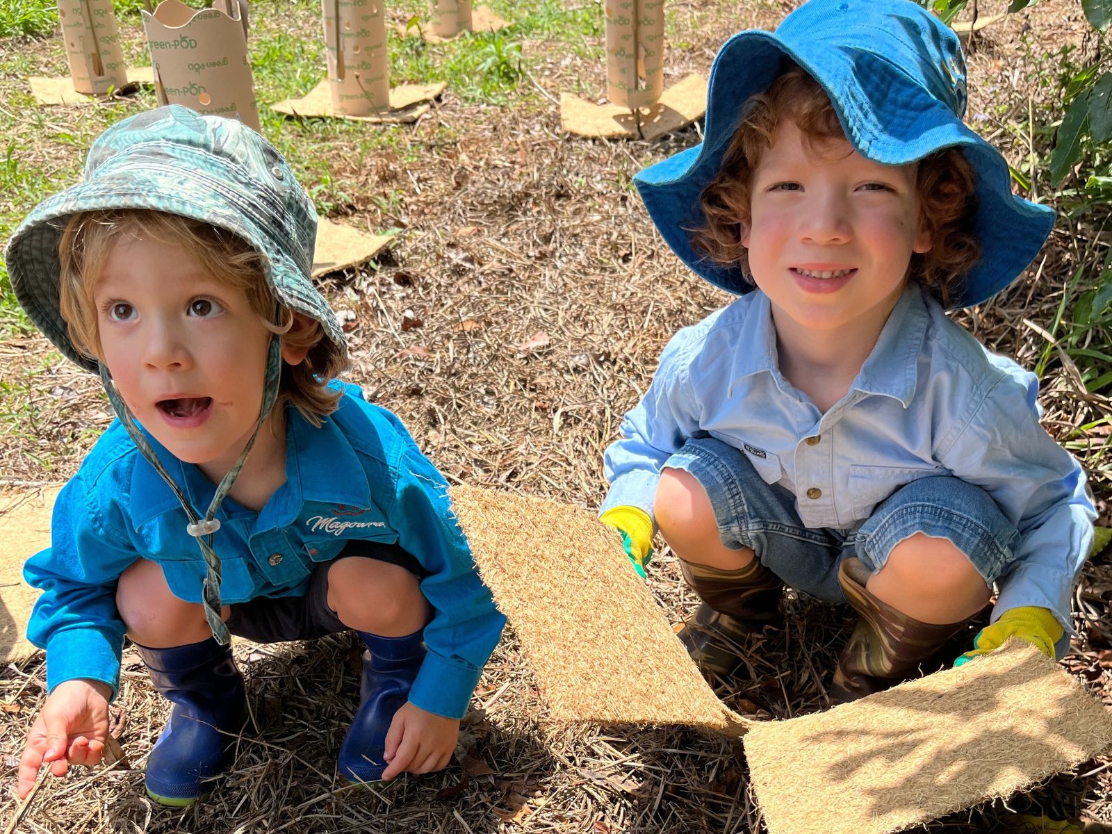 Two boys crouch, one holding plant matting.