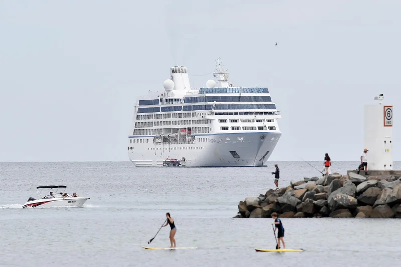 A cruise ship docked off Mooloolaba - the end of the rockwall is showing on the right, there are people on the rocks fishing. There are two stand-up paddle boarders on the water at the front of the picture and a small boat on the water on the left.