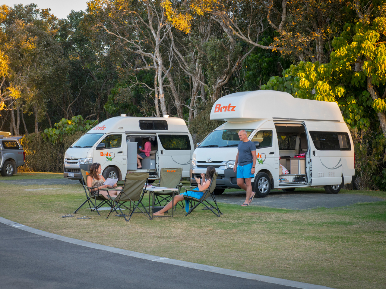 Two camper vans in the background, with two people sitting in camping chairs in front
