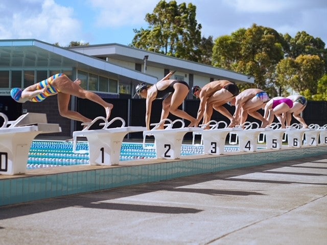Six professional swimmers on starting blocks around an outdoor swimming pool jumping in to start a race. 